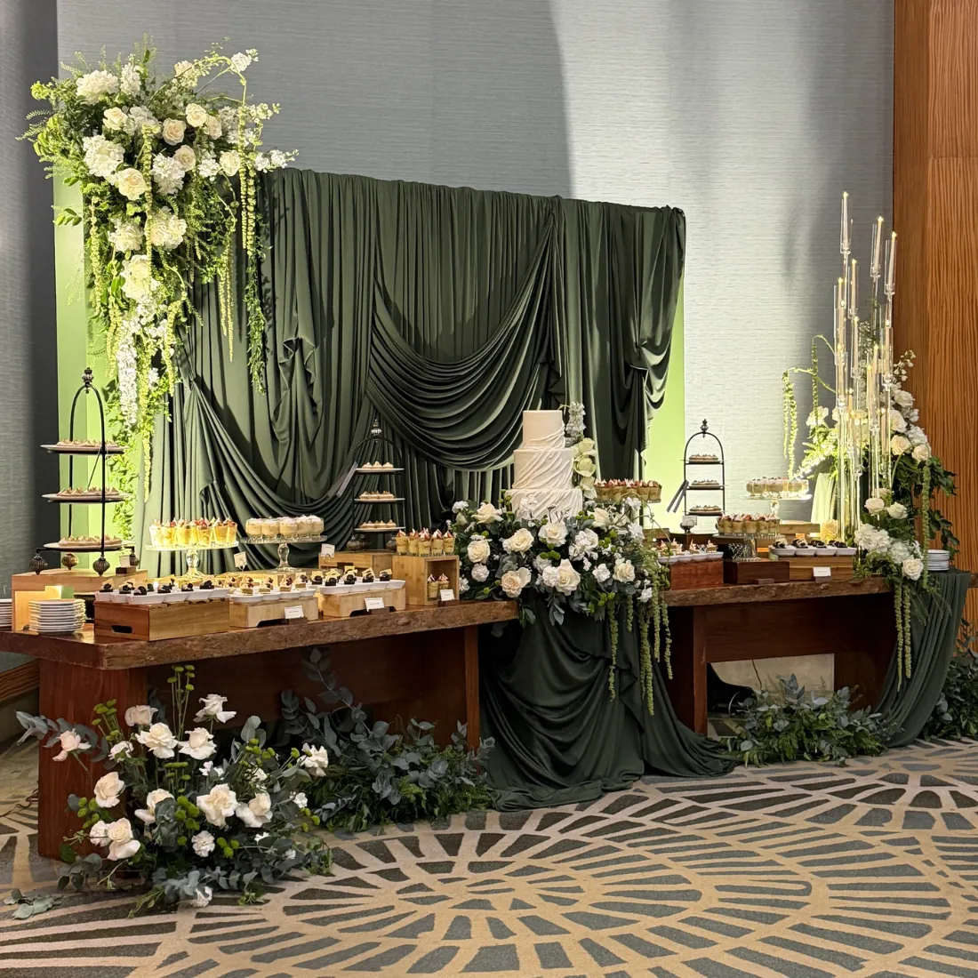 Mesa de postres con fondo de telas verdes y flores blancas